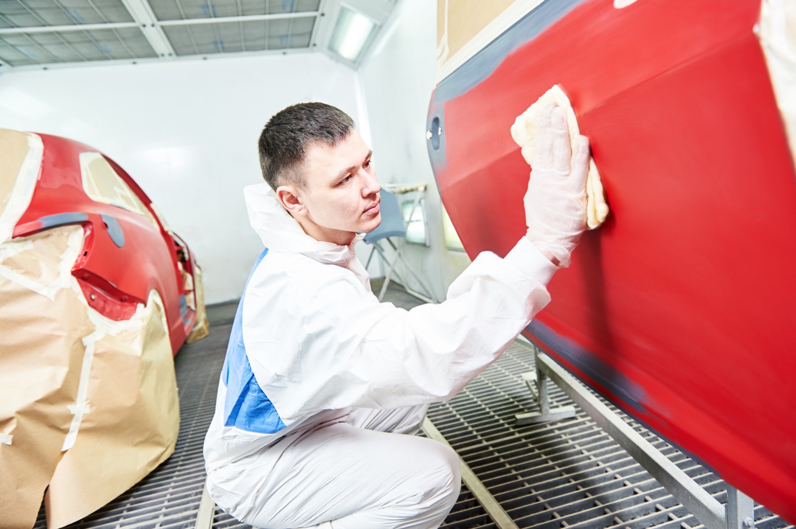 Young male paint finisher in overalls working on a red car door. The rest of the car is visible in the background